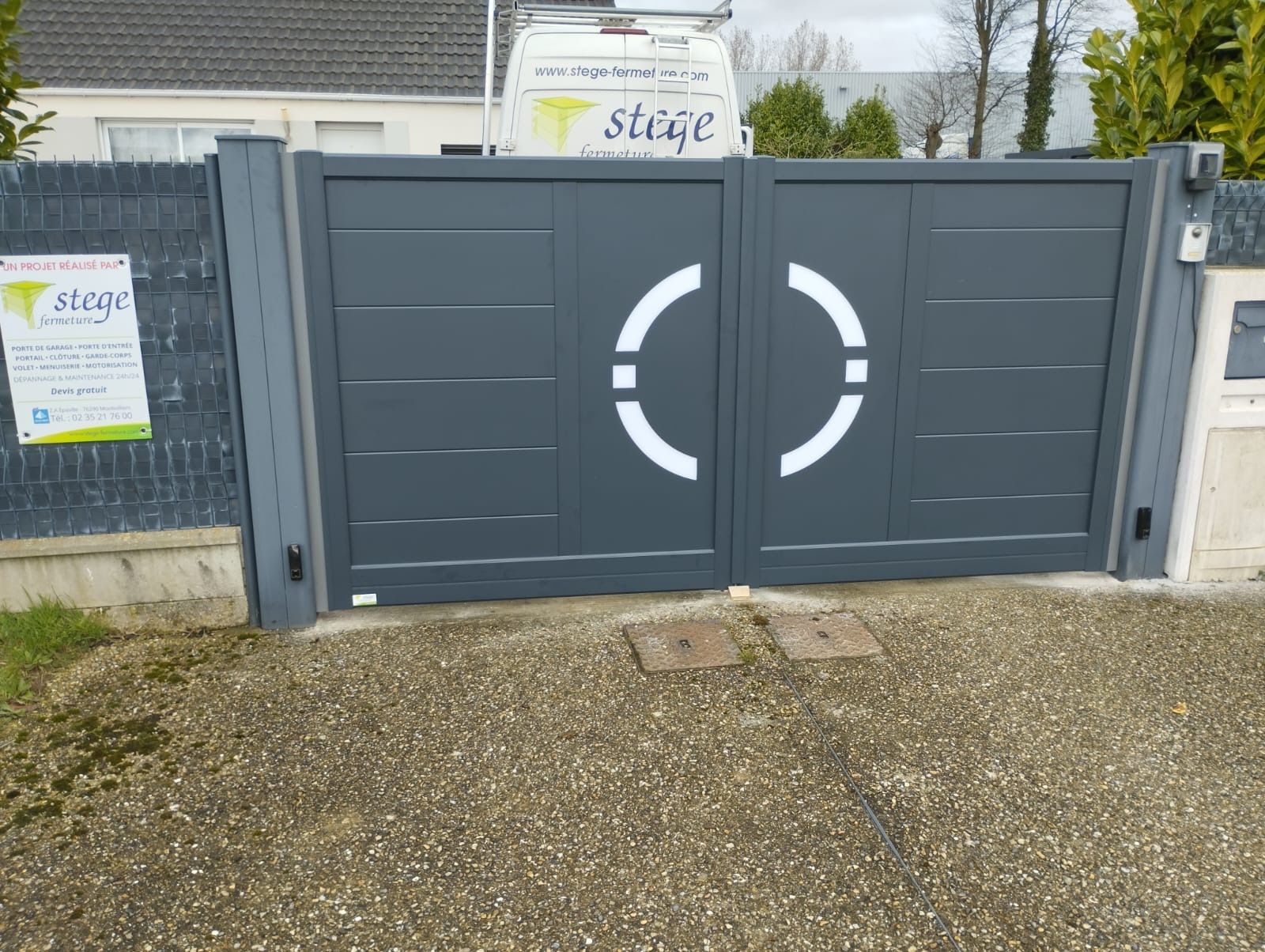 Portail métallique gris avec logo circulaire blanc, placé devant une entrée clôturée et un panneau.