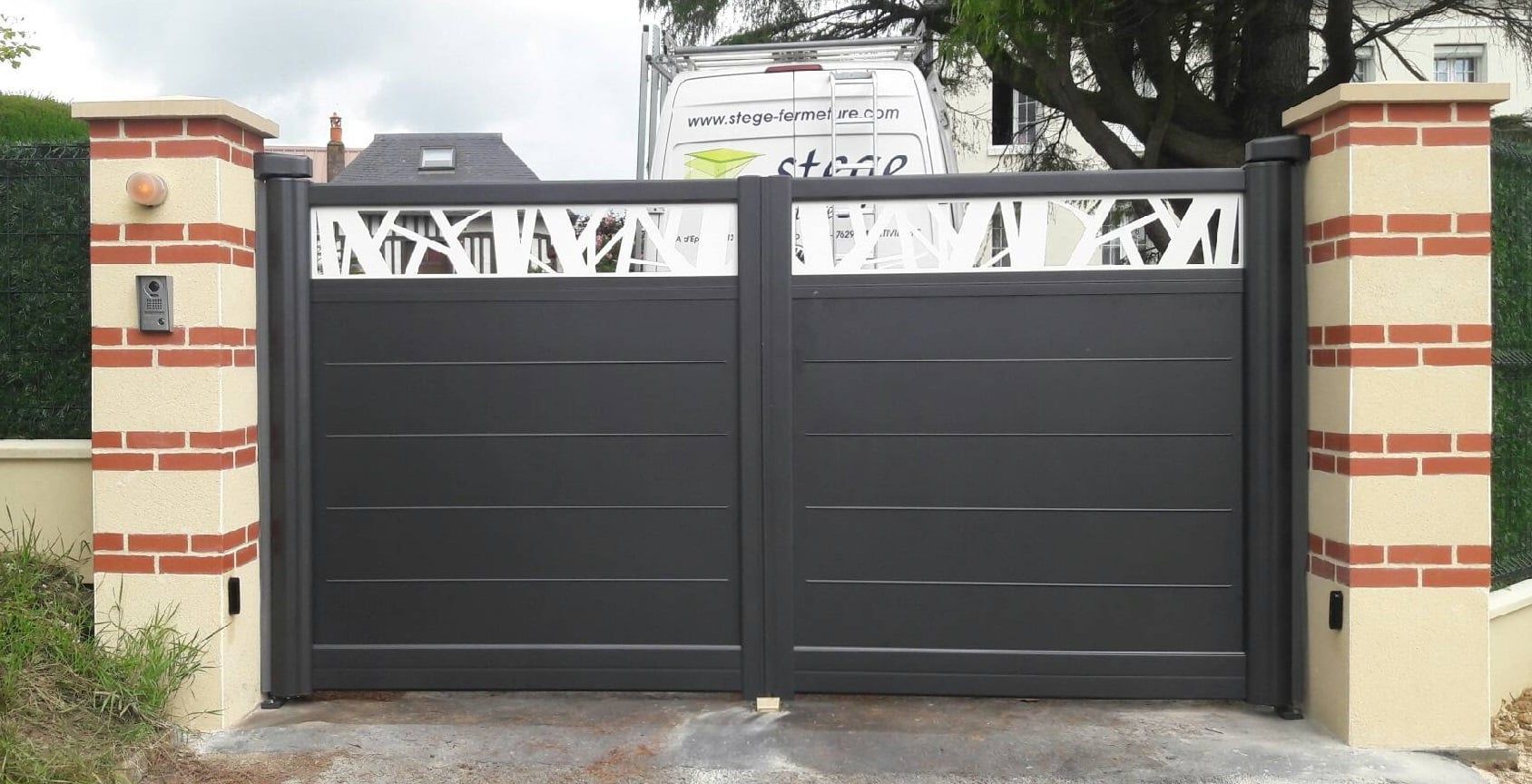 Portail d'allée fermé en métal gris foncé avec panneaux décoratifs blancs, flanqué de piliers en briques.