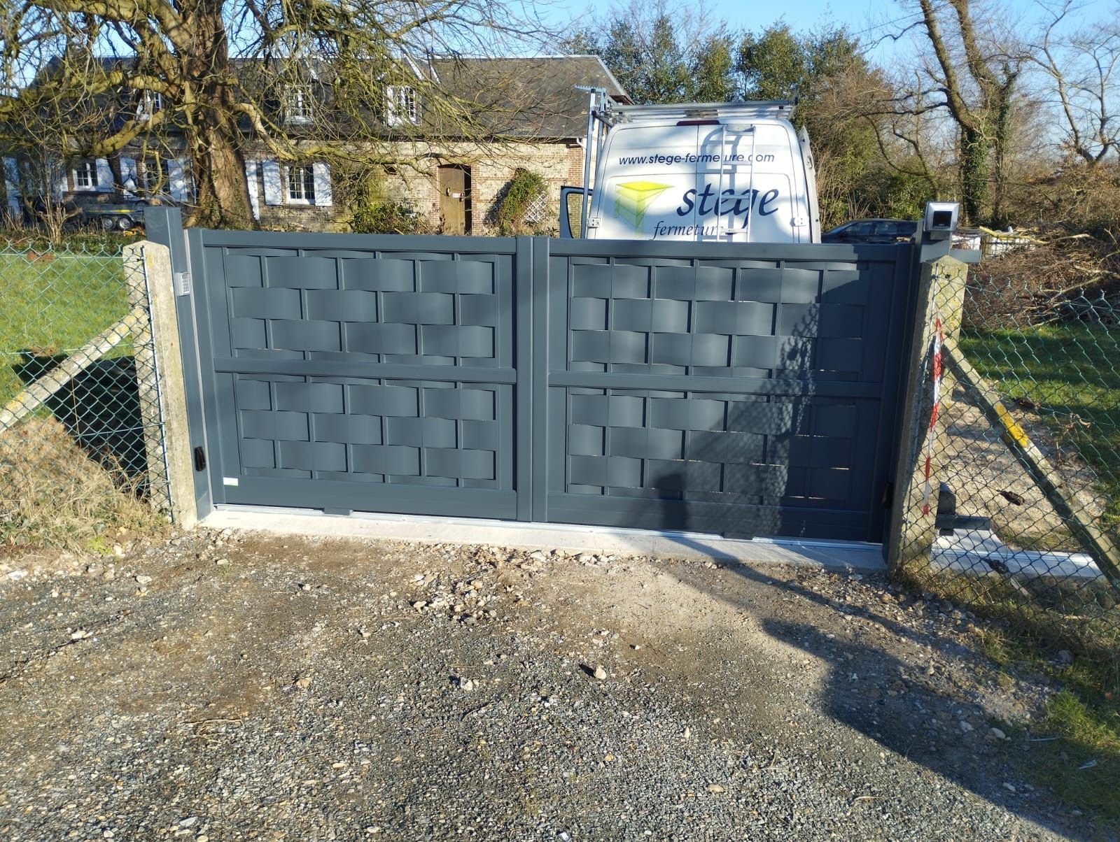 Un portail double gris foncé marque l'entrée d'une allée, avec une clôture en grillage et une cuve blanche derrière.