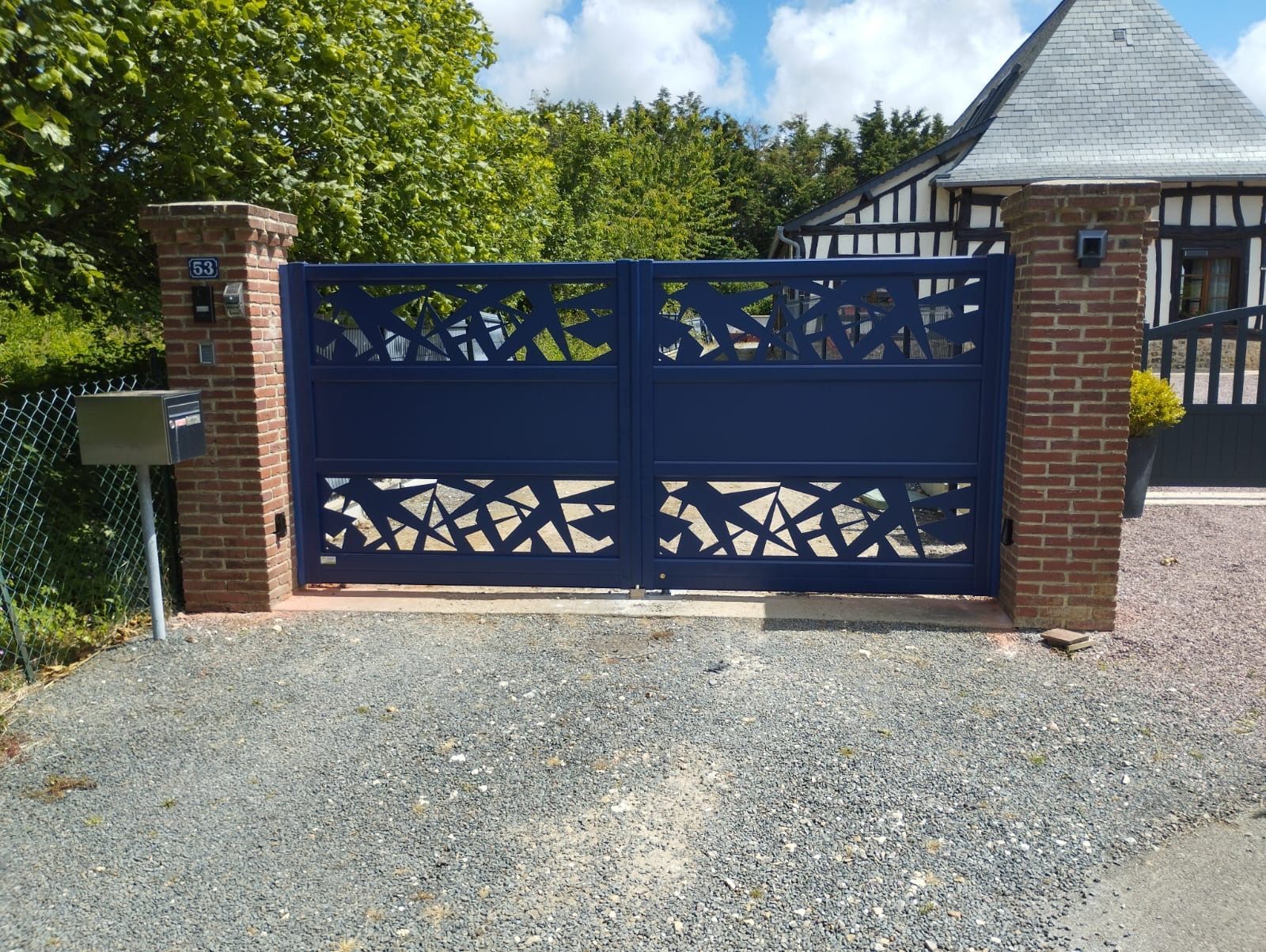 Portail ornemental bleu entre des piliers de brique à une entrée de gravier avec une maison et des arbres derrière.