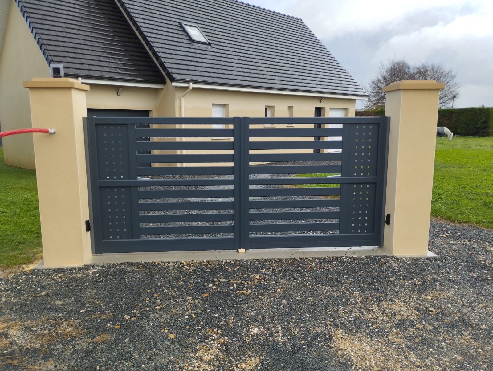 Portail coulissant gris foncé devant une maison couleur crème avec un toit de tuiles et une allée en gravier.