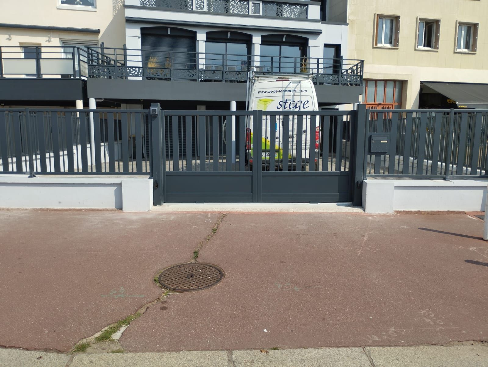 Un portail en métal noir se dresse devant un bâtiment, derrière lequel une camionnette blanche est garée sur le trottoir.