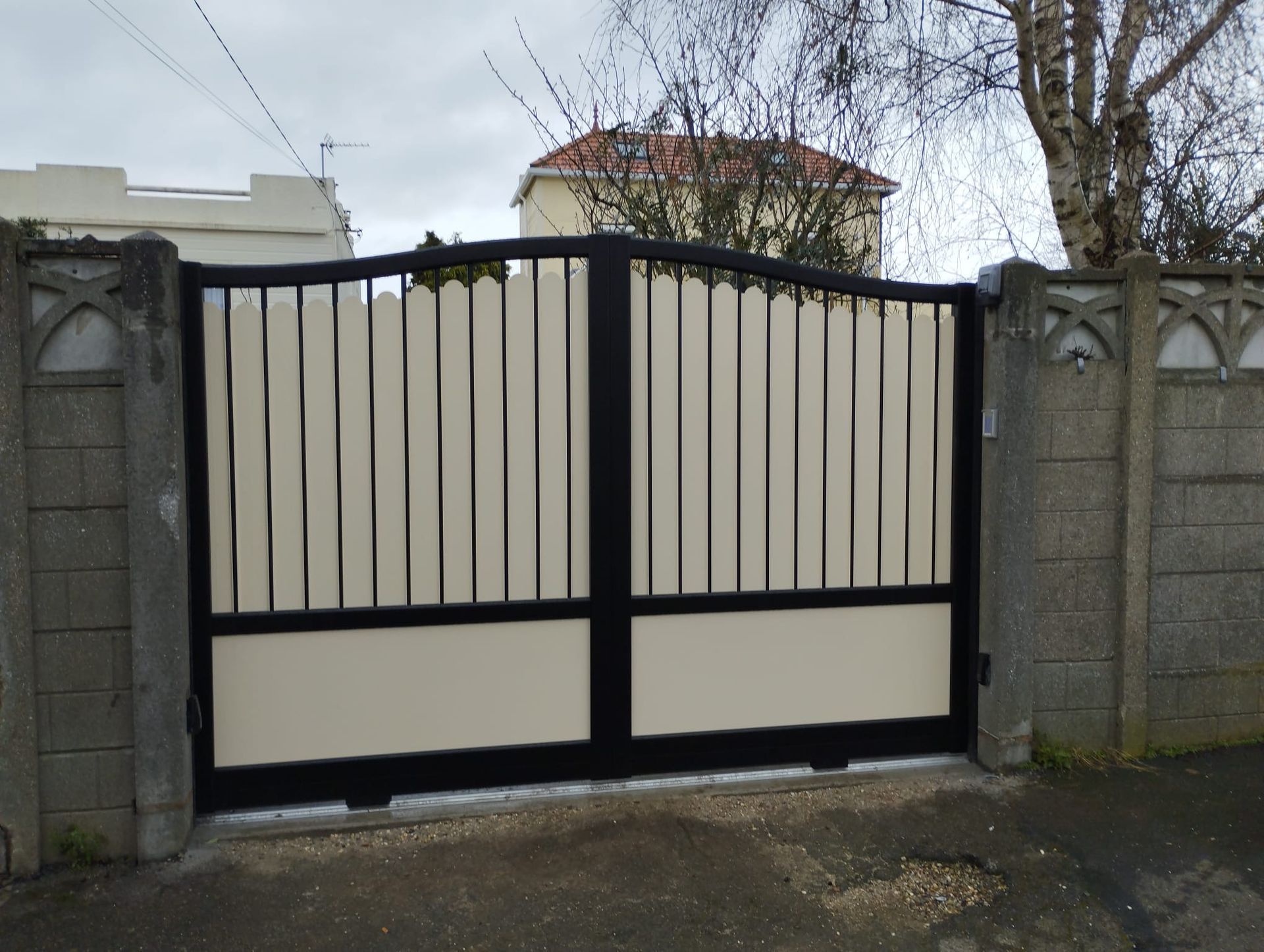 Portail métallique beige et noir fermé entre des piliers en pierre devant une maison