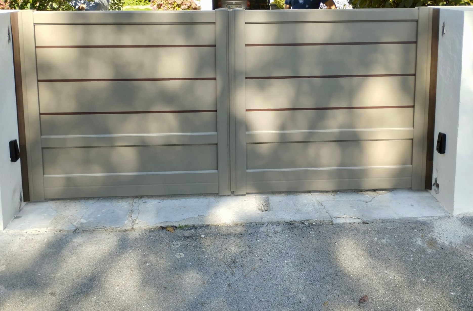 Portail métallique beige fermé dans une allée en béton, baignée de soleil et ombragée par les arbres.