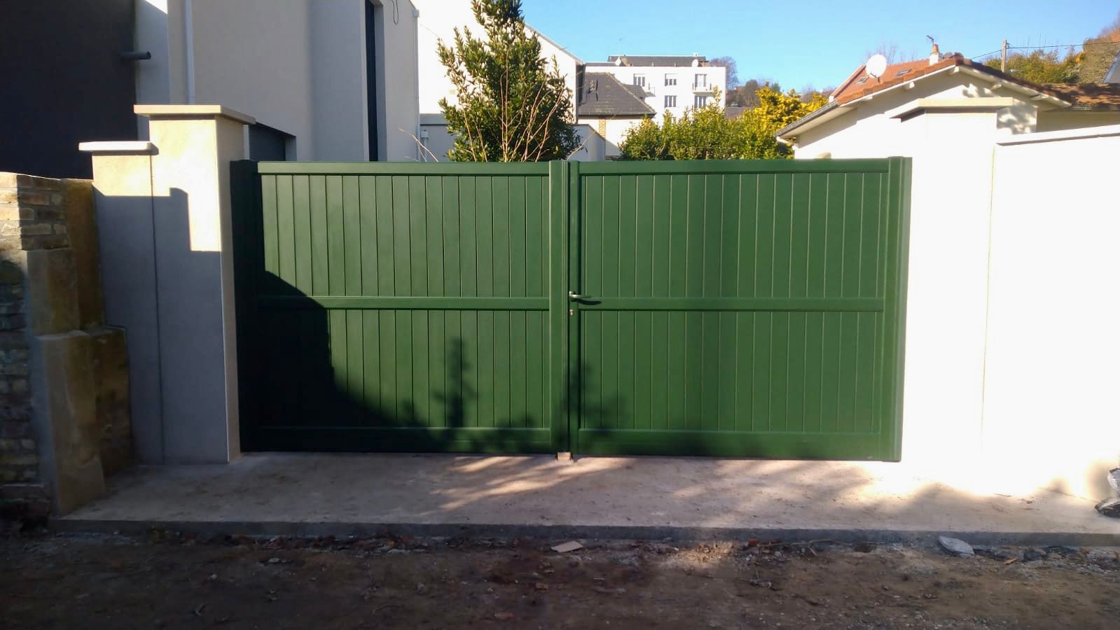 Portail double vert dans un mur blanc à l'entrée d'une allée résidentielle, avec des bâtiments en arrière-plan.