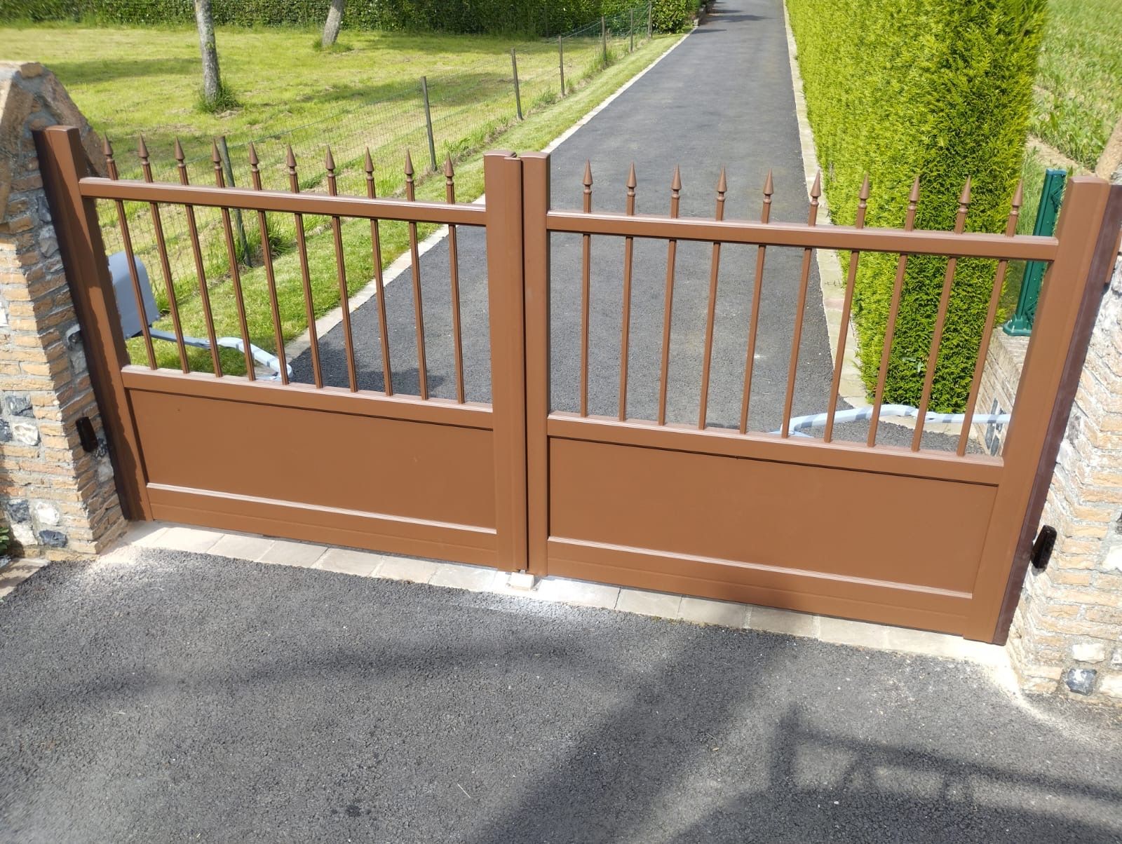 Un portail en bois brun barrait une allée, avec des barreaux métalliques et un chemin bordé de haies au-delà.