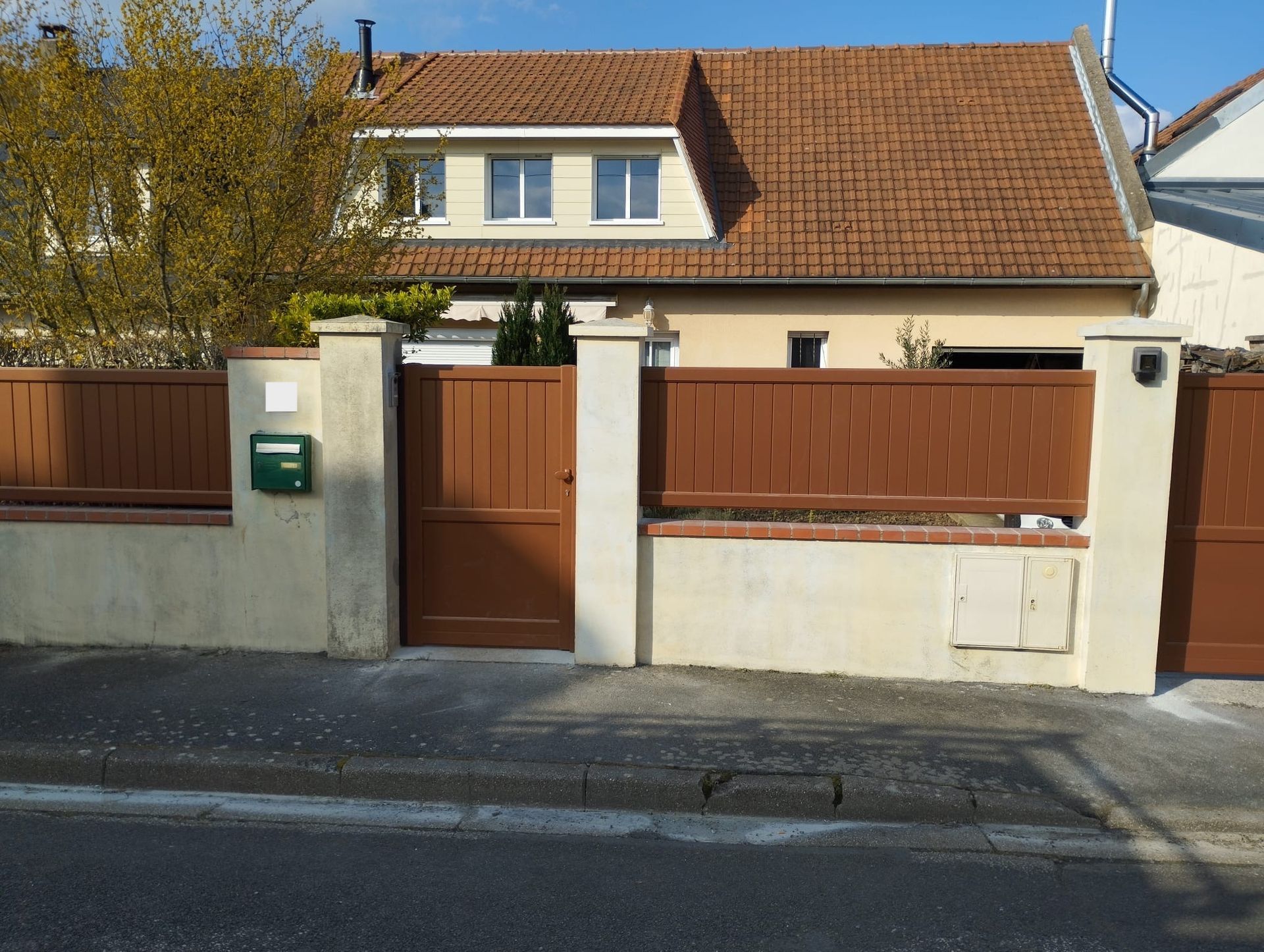 Maison de banlieue aux murs couleur crème, portail marron et toit de tuiles, derrière une clôture de rue.