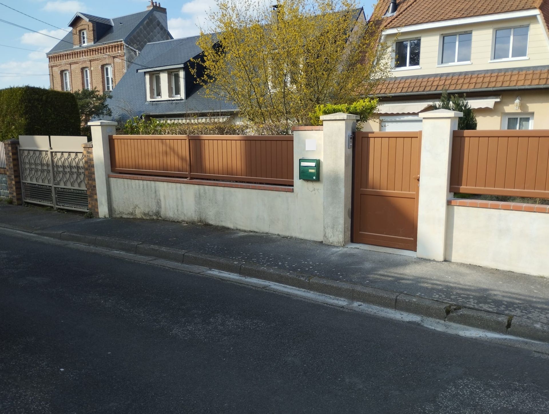 Rue résidentielle avec un muret, un portail en bois et des maisons derrière, par une journée ensoleillée