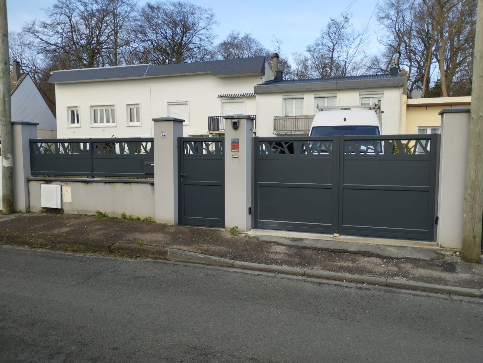 Maison moderne derrière un portail gris et un mur blanc, avec des arbres dénudés et une rue en face