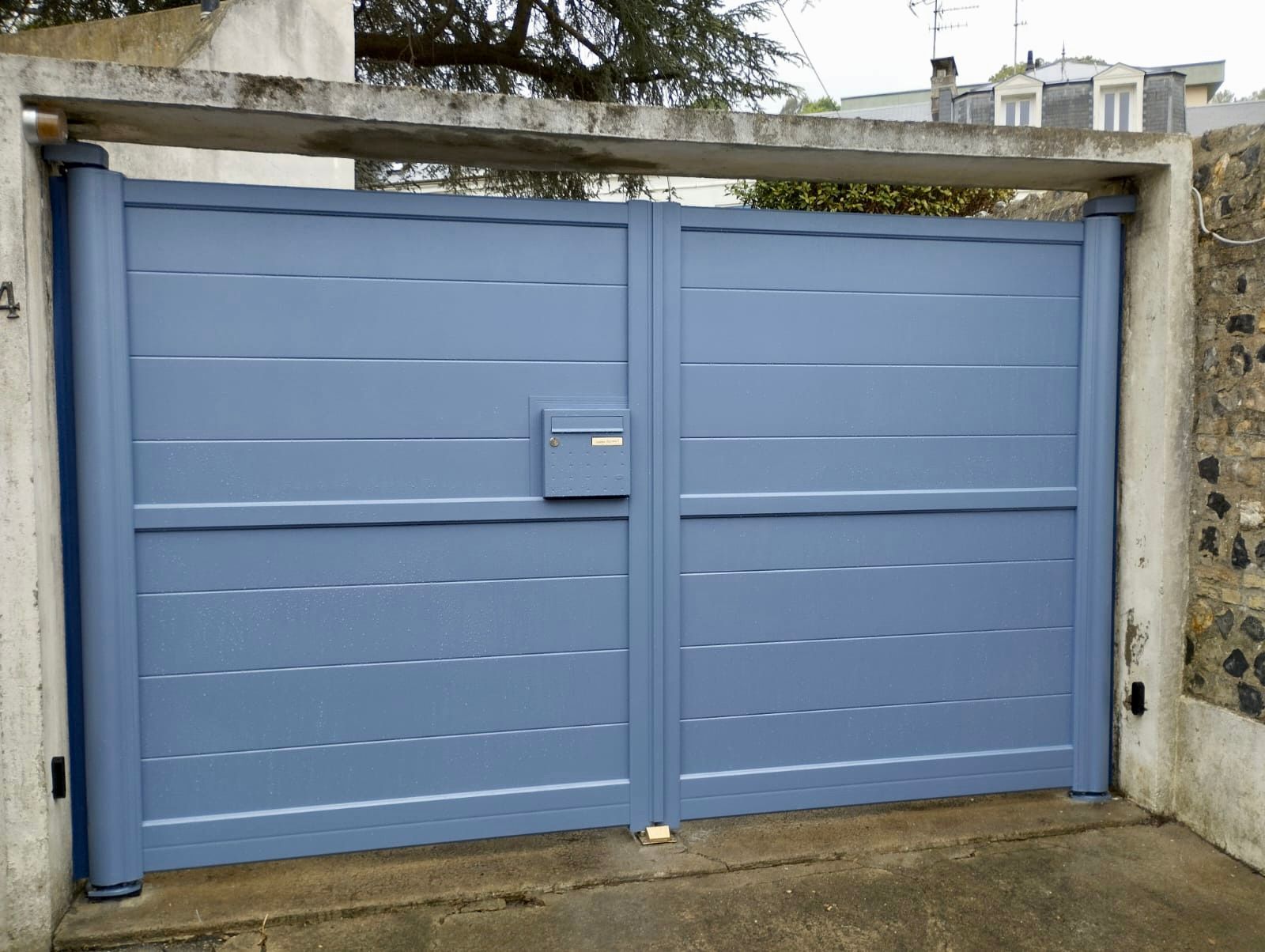 Portail double en métal bleu avec une petite boîte aux lettres en son centre, installé dans une entrée de mur en pierre