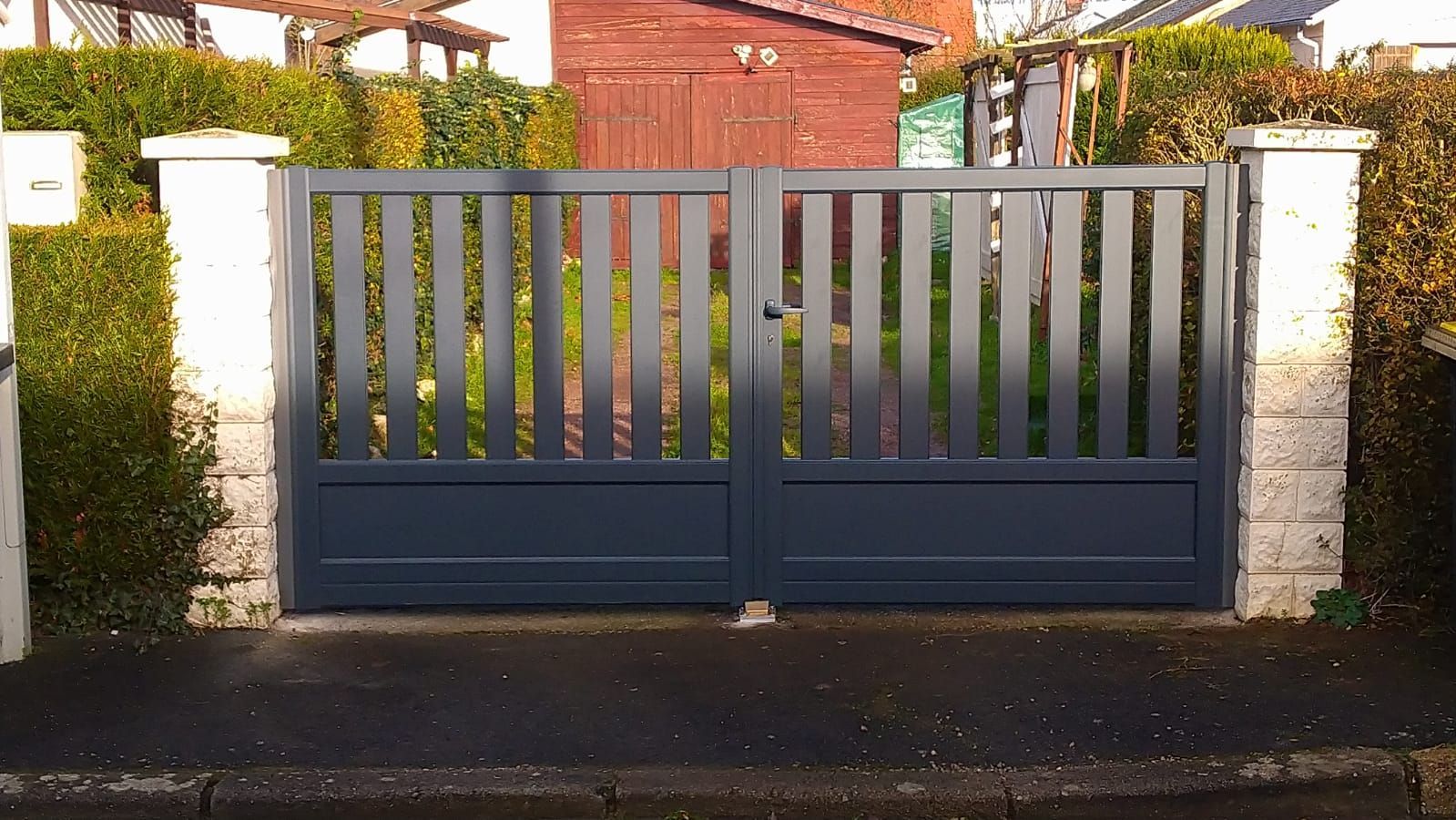 Portail d'allée en métal bleu foncé avec piliers en pierre blanche devant une maison.