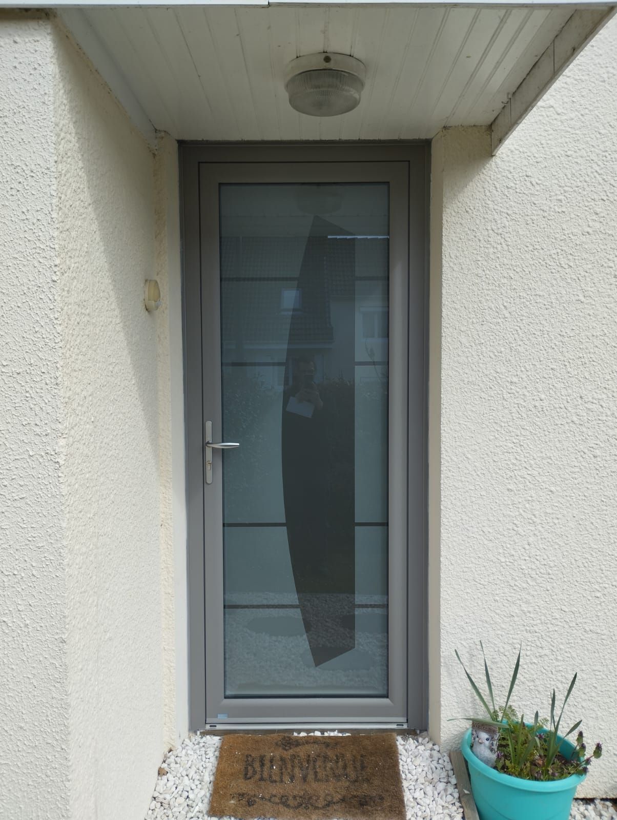 Entrée principale avec porte grise, panneau de verre dépoli, paillasson et éclairage extérieur sous une alcôve couverte.