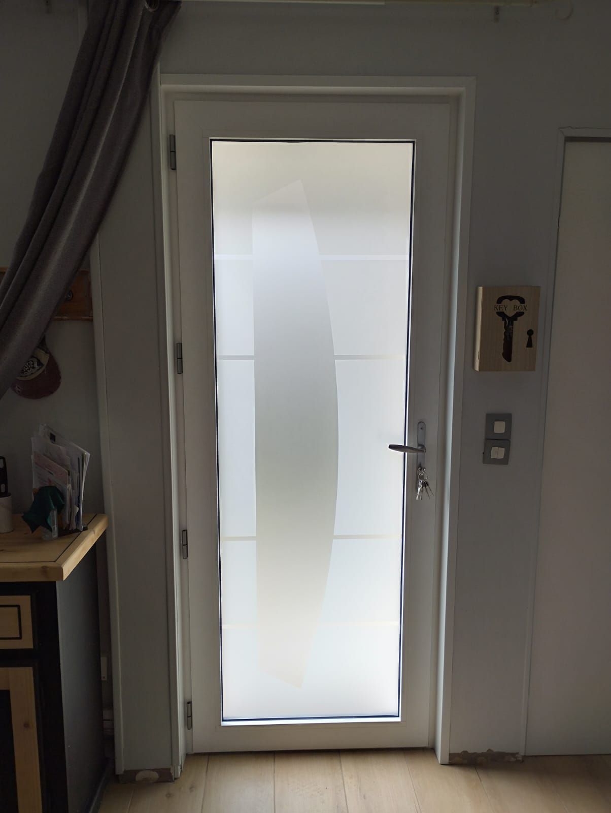 Porte d'entrée avec panneaux de verre dépoli et poignée noire donnant sur un couloir, avec un rideau et une boîte à clés à proximité.