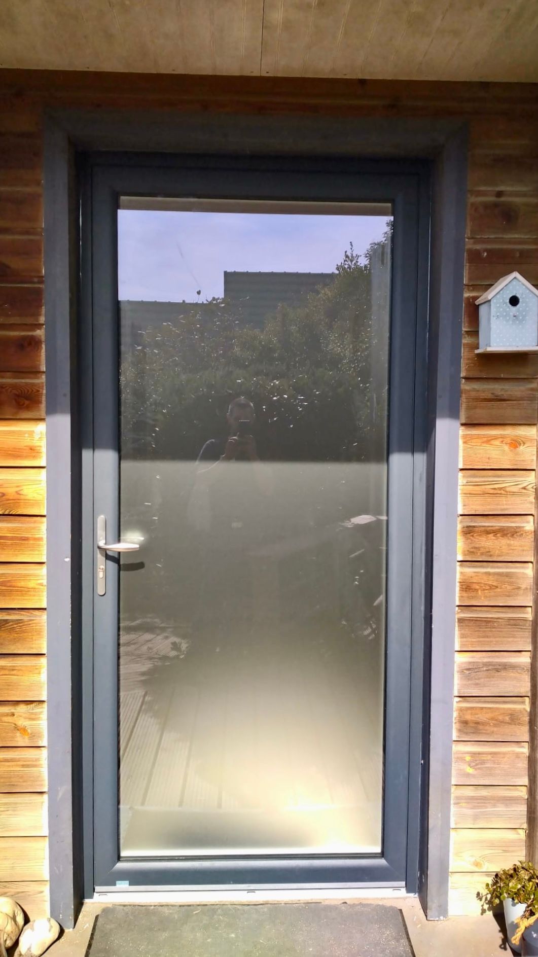 Porte d'entrée avec panneau en verre dépoli et poignée argentée, encadrée par un bardage en bois.