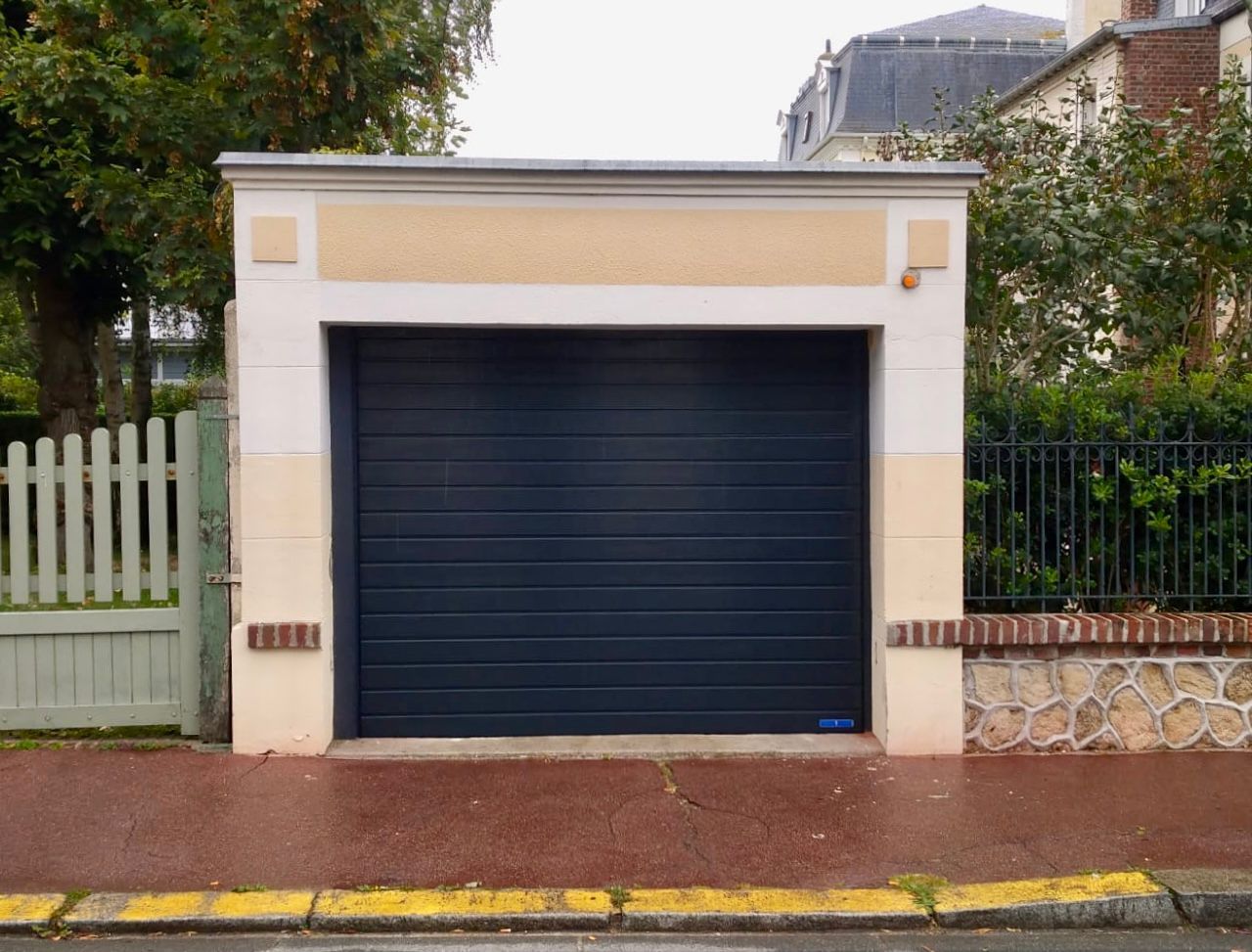 Garage blanc avec porte bleu foncé dans une rue résidentielle, flanqué d'un portail et d'un mur en pierre