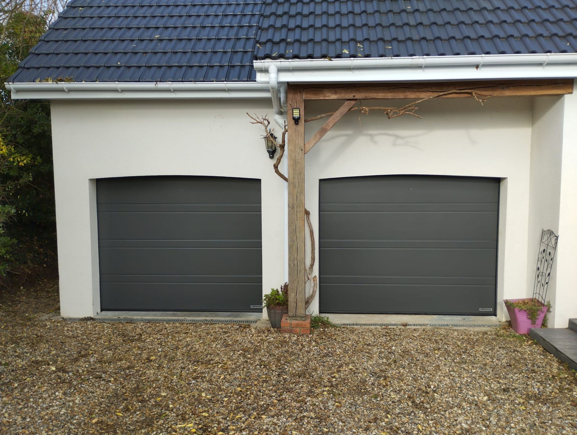 Deux portes de garage gris foncé sur une maison de couleur claire, avec du gravier devant et un tronc d'arbre entre elles