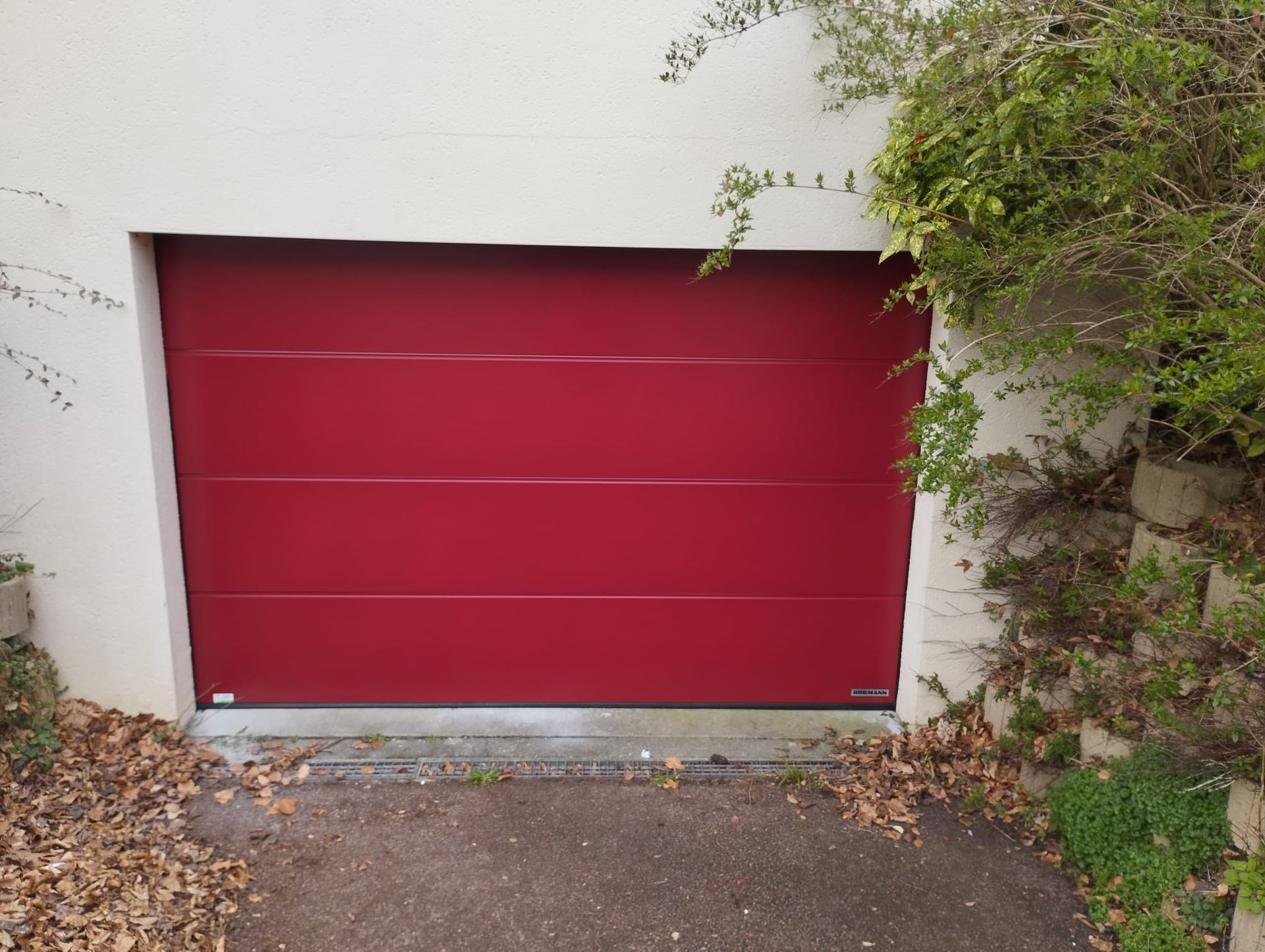 Une porte de garage rouge encastrée dans un mur blanc, entourée de feuilles et de verdure.