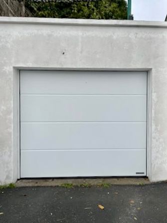 Porte de garage blanche encastrée dans un mur de béton gris, à côté d'une allée pavée.