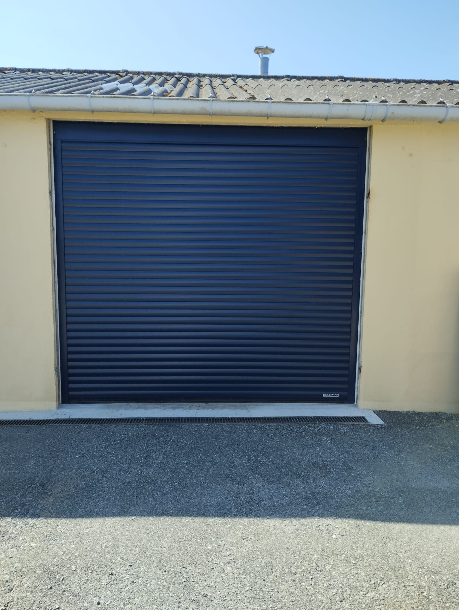 Porte de garage sectionnelle bleue fermée sur un bâtiment beige avec une allée en gravier.