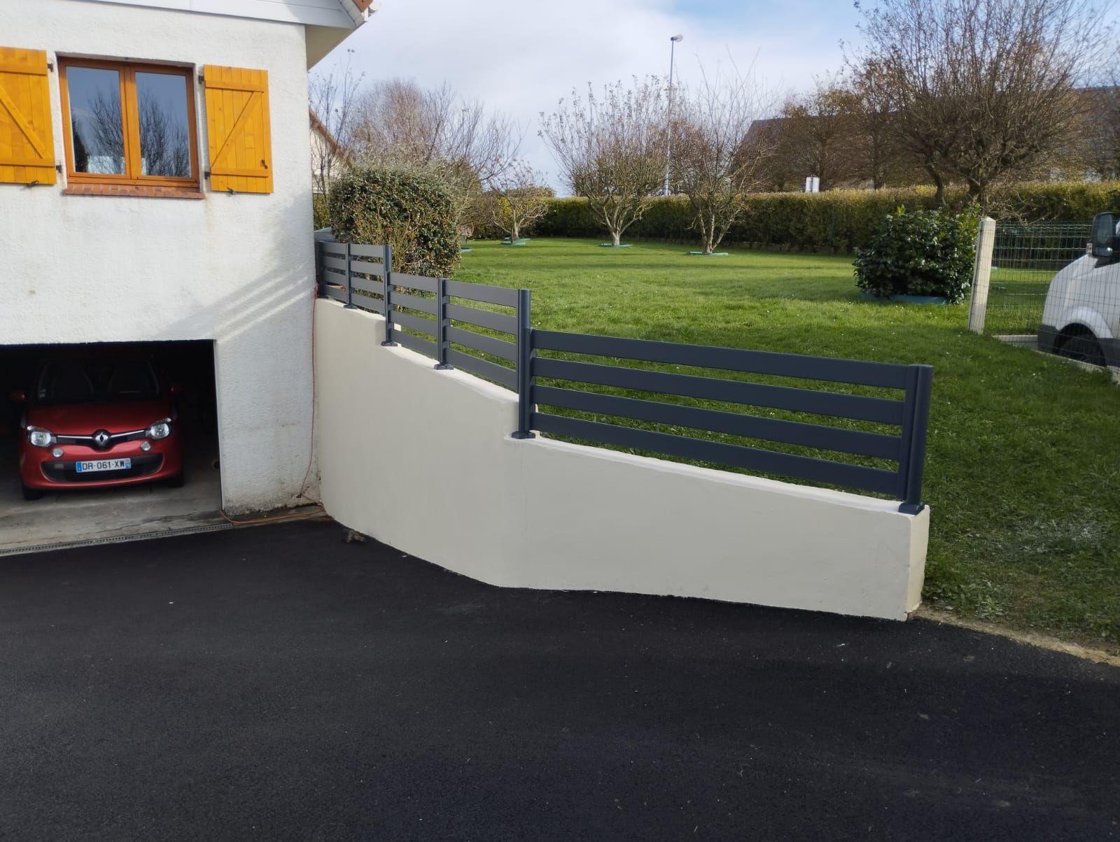 Allée de maison avec une voiture rouge dans un garage, mur blanc, clôture grise et jardin gazonné