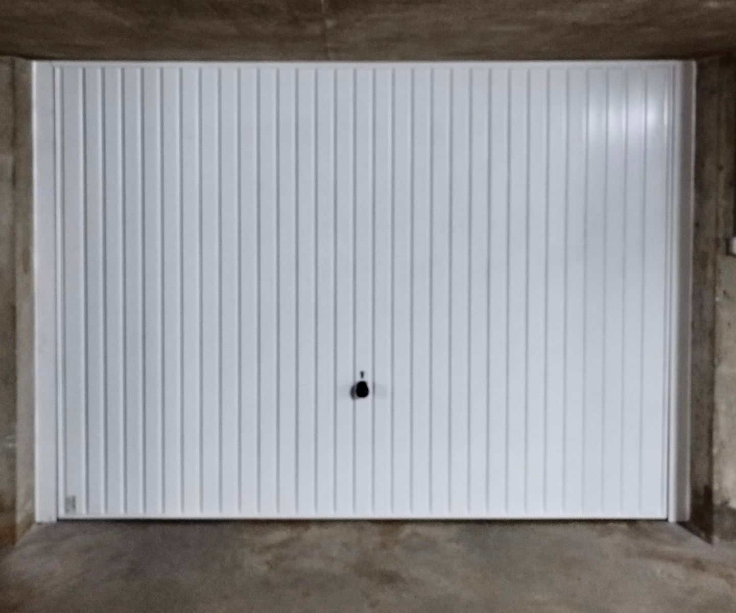 Porte de garage blanche à panneaux verticaux et poignée noire dans une ouverture sur un mur en béton