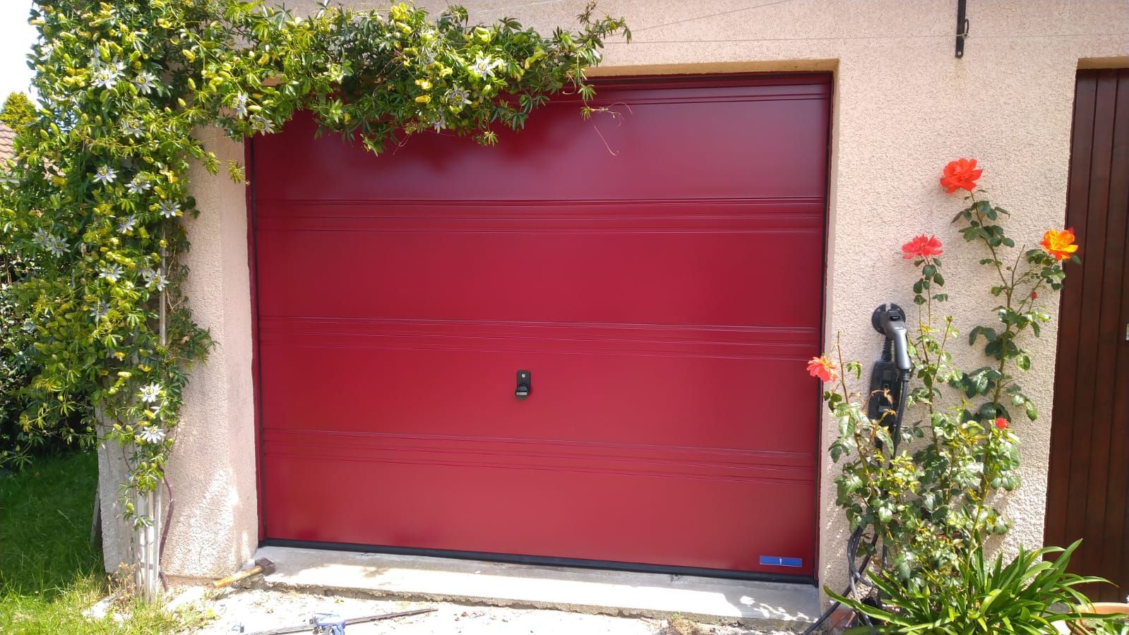 Porte de garage rouge encadrée de vignes vertes et de fleurs sur un mur en stuc