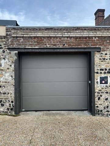 Porte de garage grise fermée dans un mur de briques et de pierres, avec une allée de gravier.