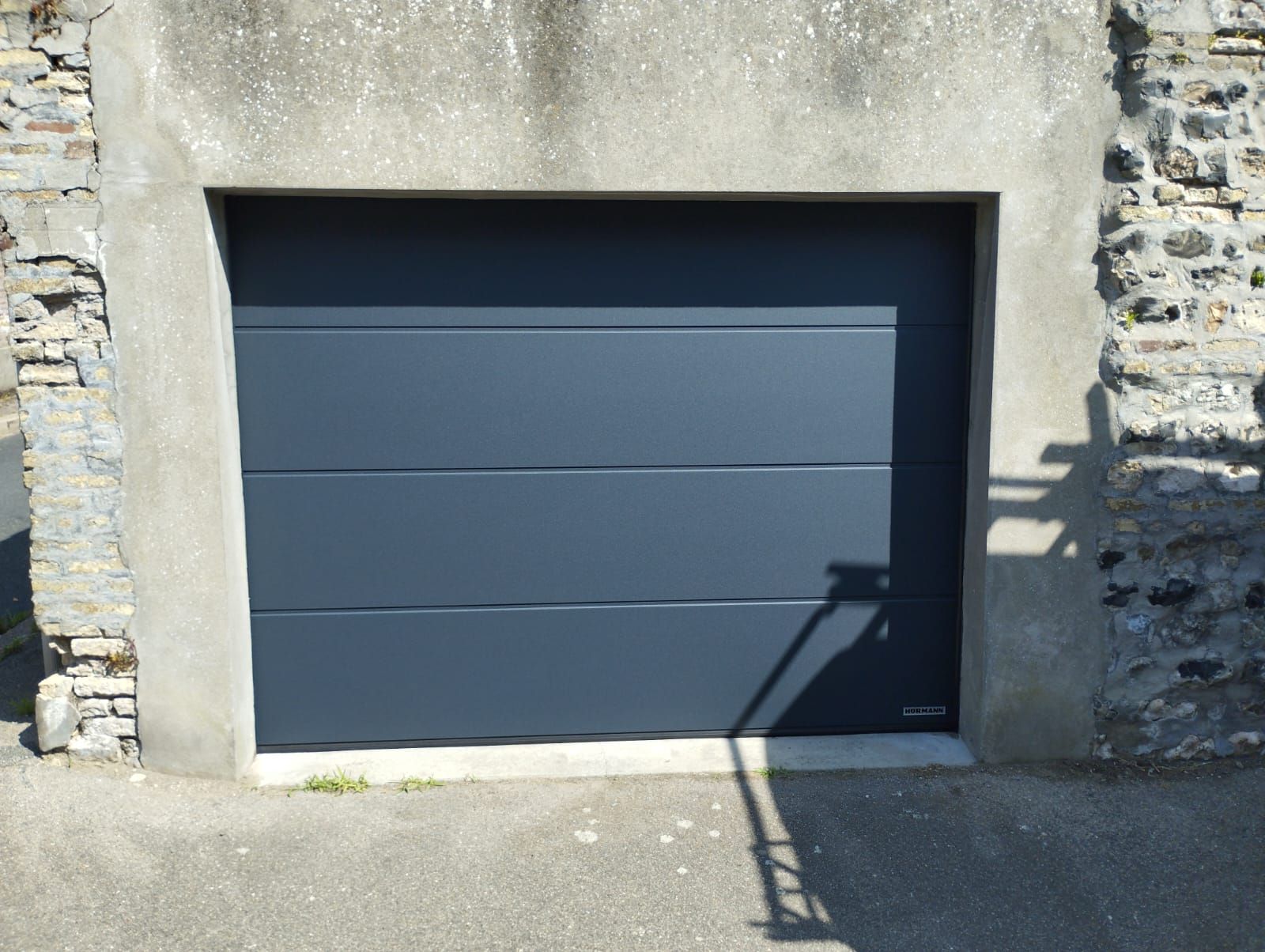 Porte de garage gris foncé encastrée dans un mur de pierre avec une petite rampe et des ombres latérales marquées.