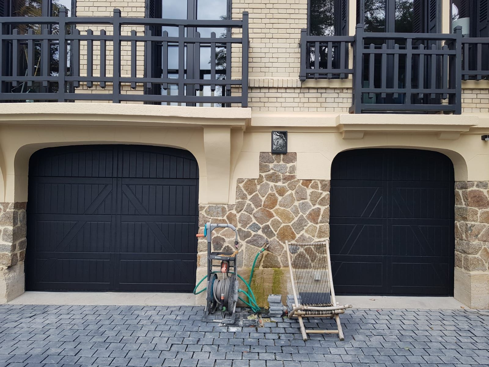 Deux portes de garage noires sous un balcon, avec façade en pierre et une petite échelle et des outils devant