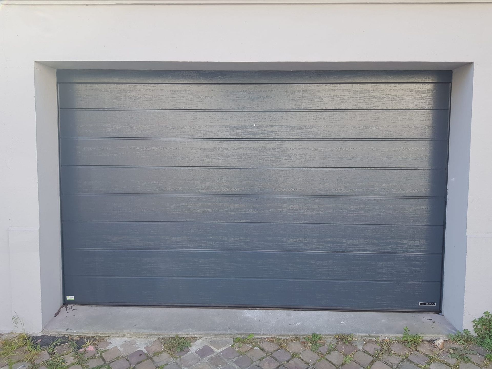 Porte de garage fermée gris foncé encastrée dans un mur blanc au-dessus d'une allée pavée