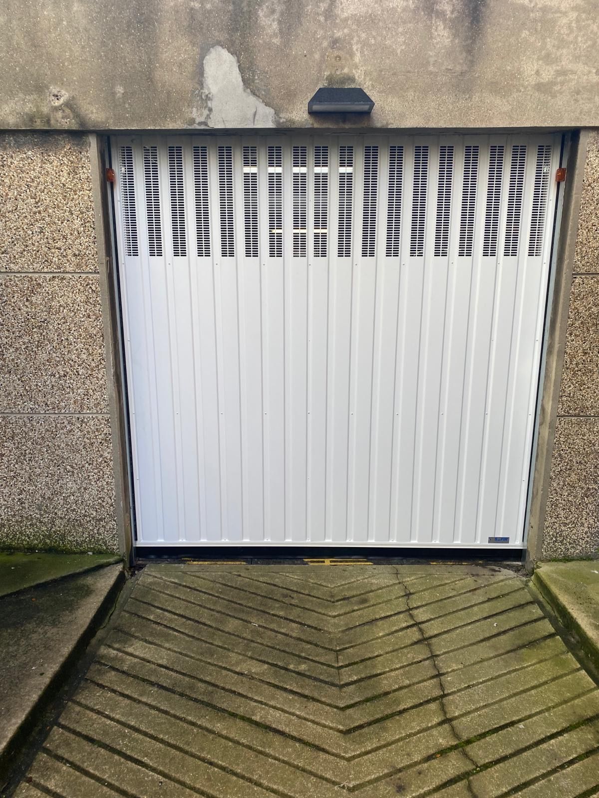 Porte de garage blanche encastrée dans un mur en béton, avec une allée à motifs devant.