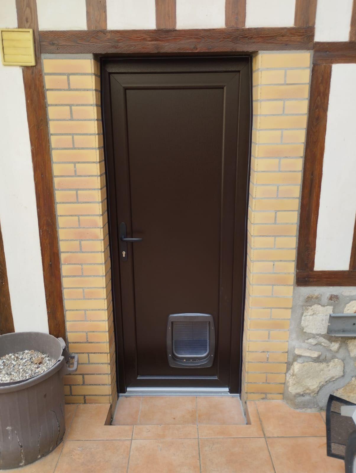 Porte marron foncé avec chatière dans une entrée en briques et à colombages, à côté d'une poubelle.