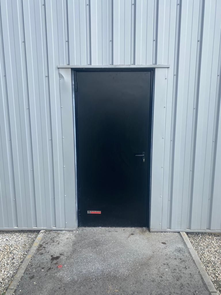 Porte noire dans un mur en tôle ondulée, avec un chemin de gravier y menant.