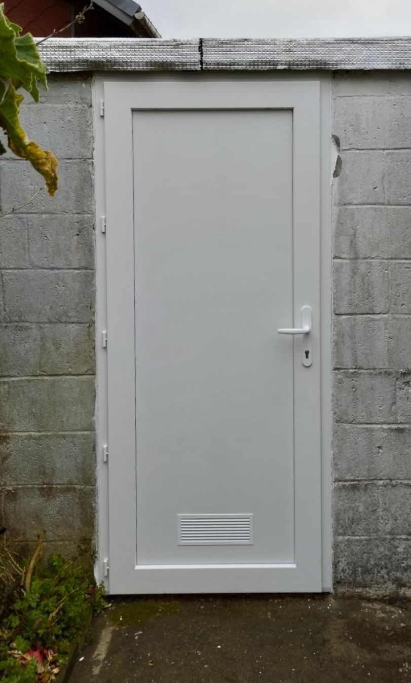 Porte extérieure blanche encastrée dans un mur en parpaings de béton gris, avec une petite grille d'aération en bas.