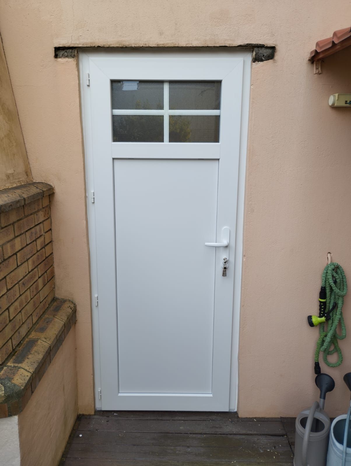 Porte extérieure blanche à petits carreaux, encastrée dans un mur beige à côté d'une marche en briques et d'un tuyau d'arrosage.