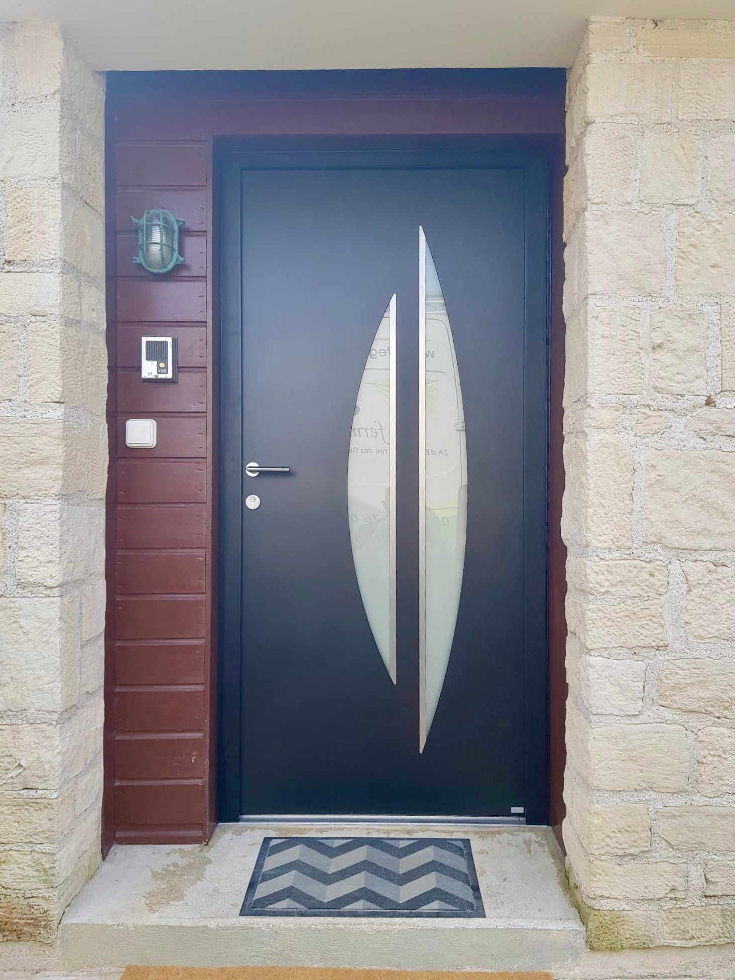 Porte d'entrée avec vitrage décoratif noir et blanc, murs en pierre et paillasson rayé.