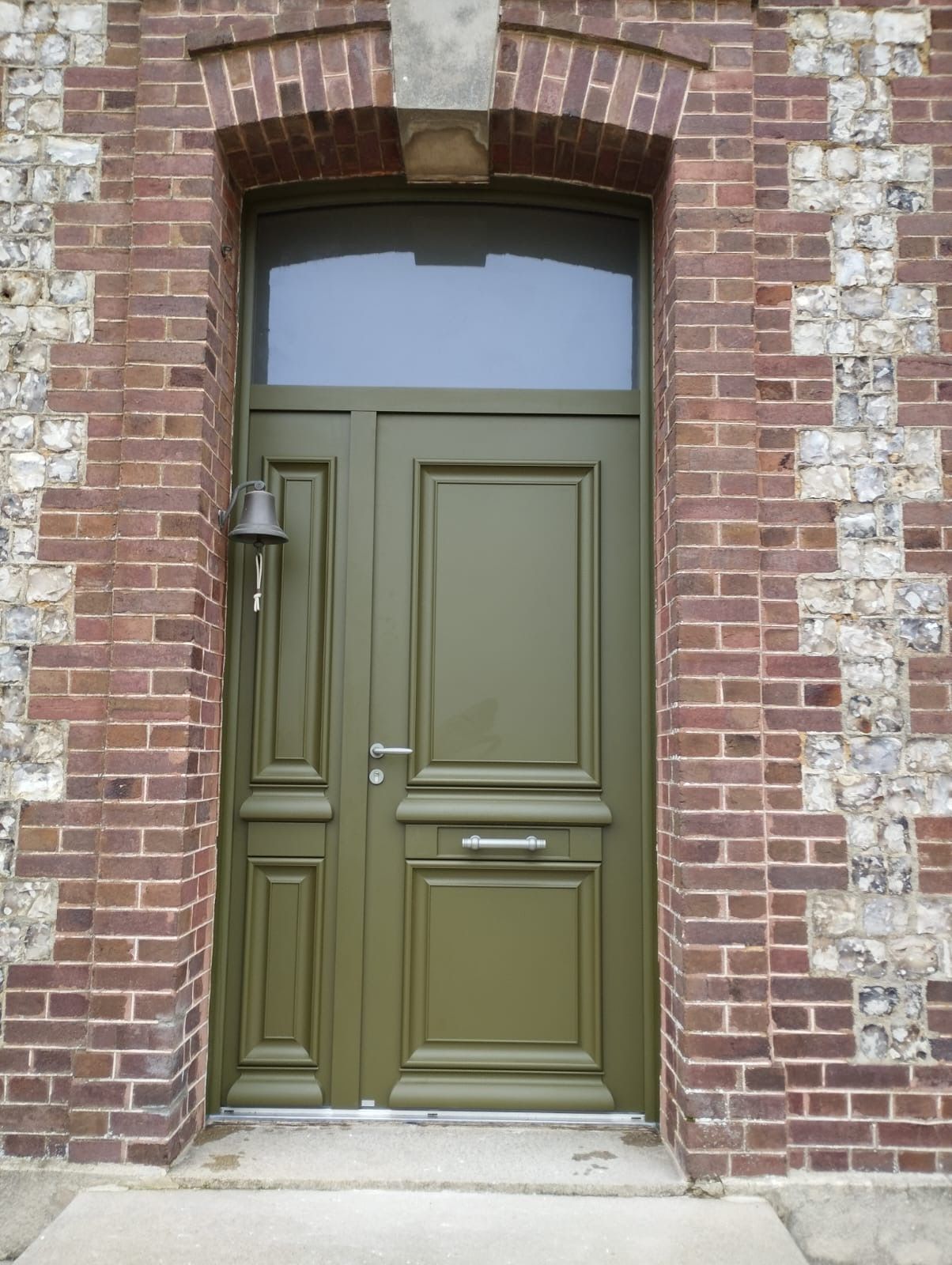 Porte double vert olive encastrée dans une arche en briques, avec une petite fente pour boîte aux lettres et une lampe sur la gauche.