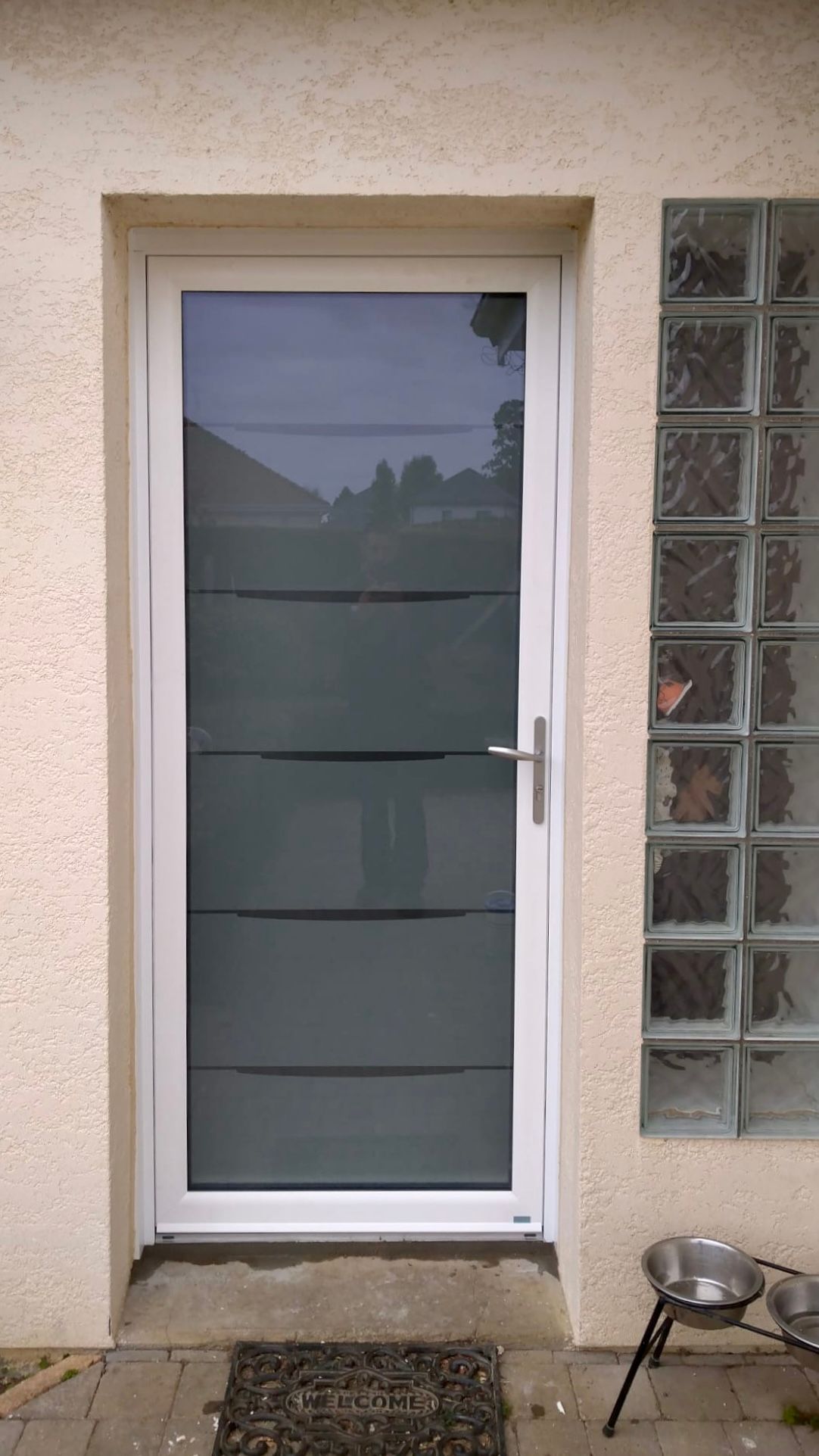 Porte d'entrée blanche avec panneaux de verre dépoli, à côté d'un mur en pavés de verre et d'un petit tabouret sur une terrasse.