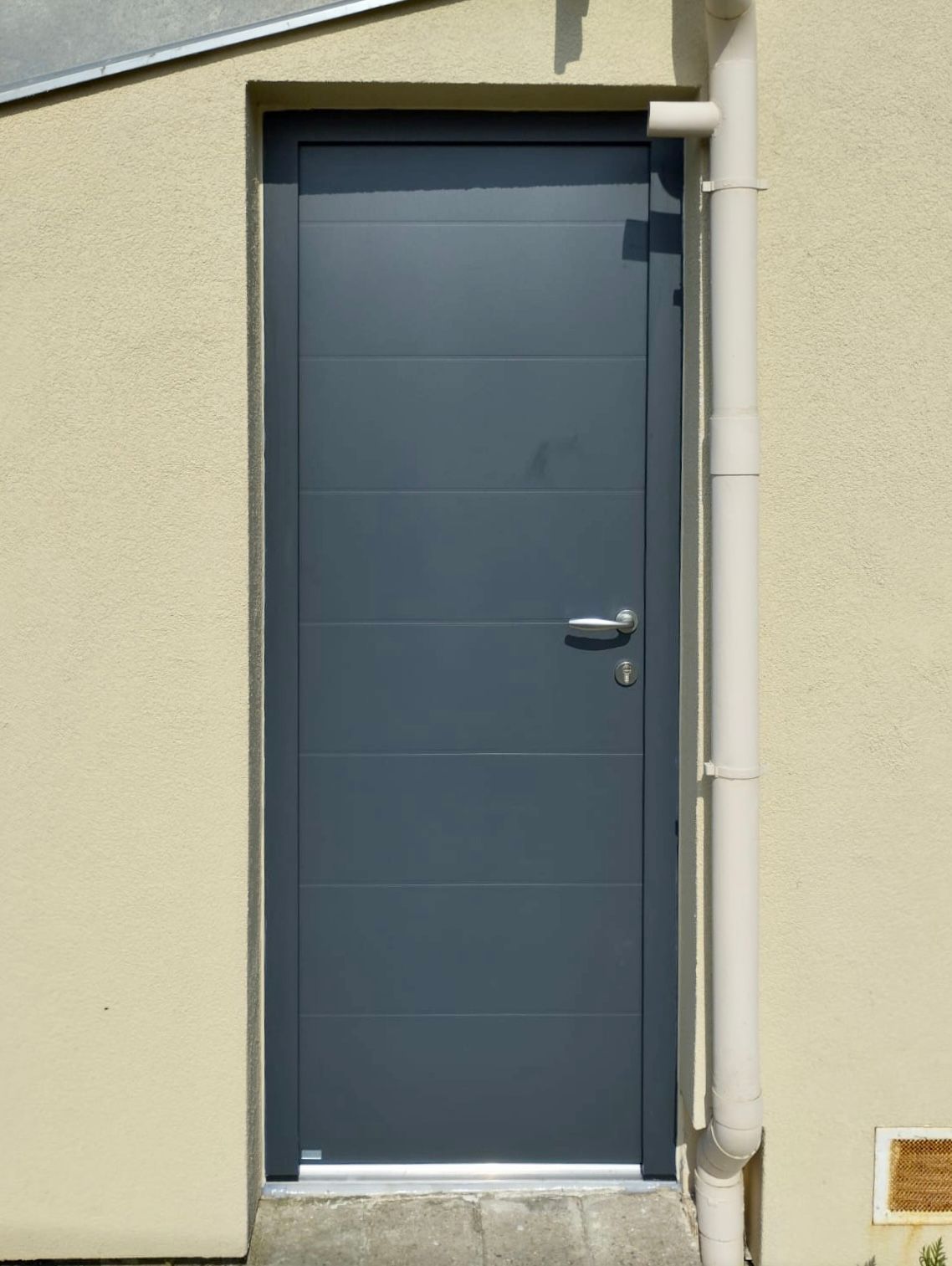 Porte extérieure en métal gris sur un mur beige avec une descente de gouttière blanche à côté.