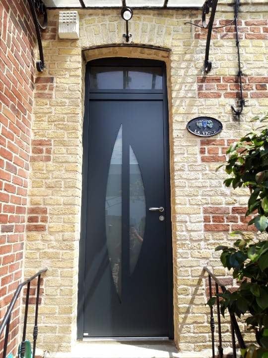 Porte d'entrée vitrée étroite dans un immeuble en briques, encadrée par une balustrade noire et de la verdure.