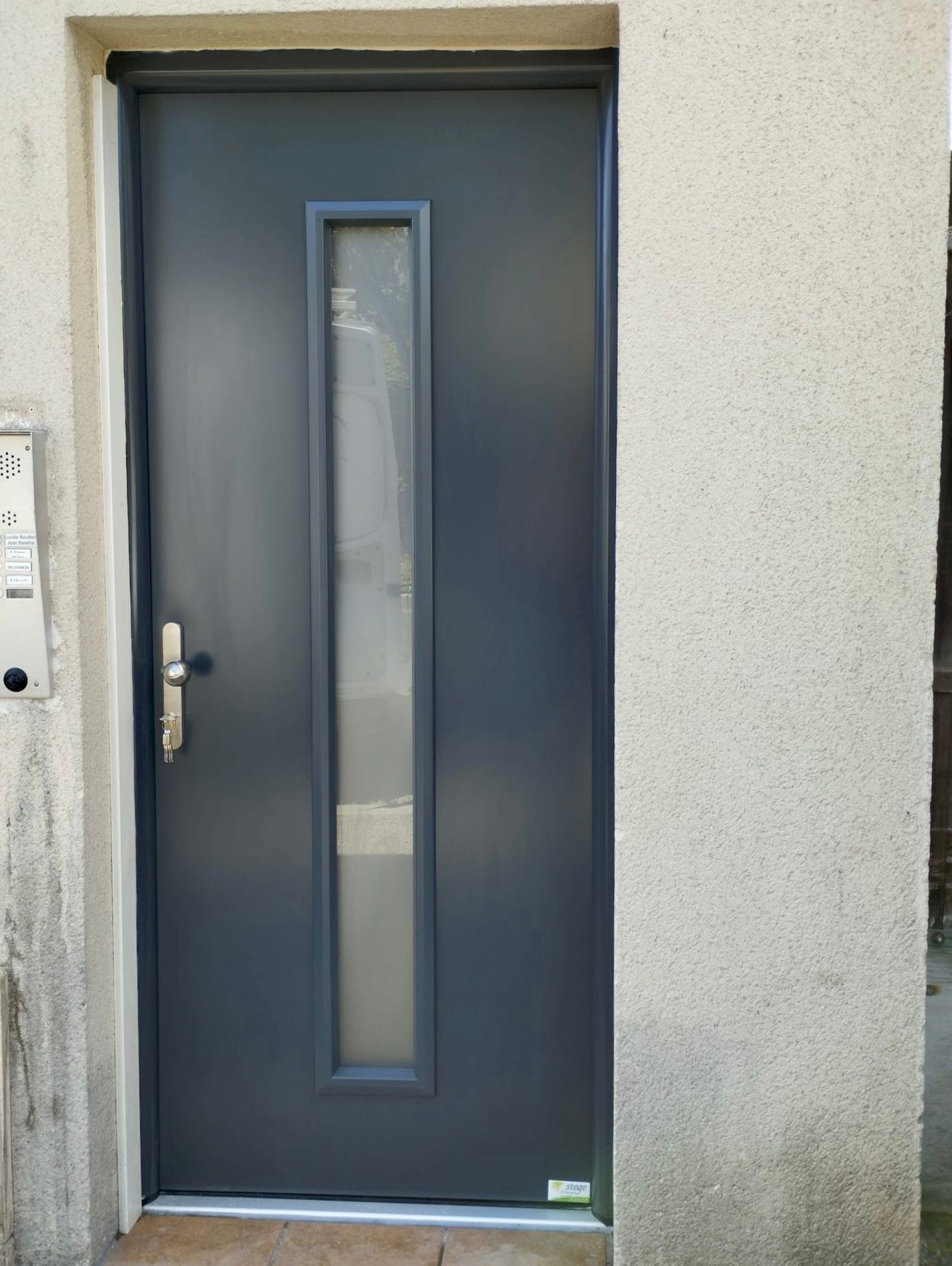 Porte d'entrée grise avec une étroite fenêtre verticale et une poignée argentée dans un mur en stuc