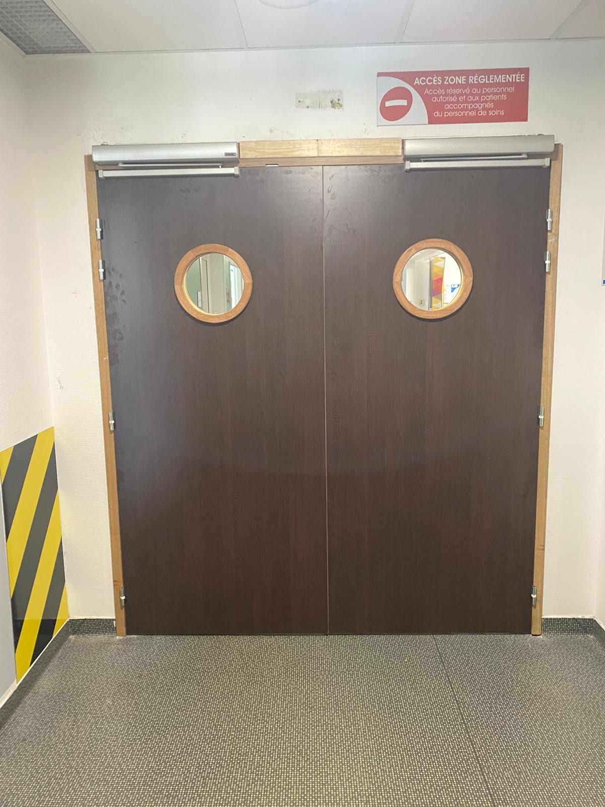 Porte double marron foncé avec fenêtres rondes dans un couloir, bordée de bandes de signalisation jaune et noire.
