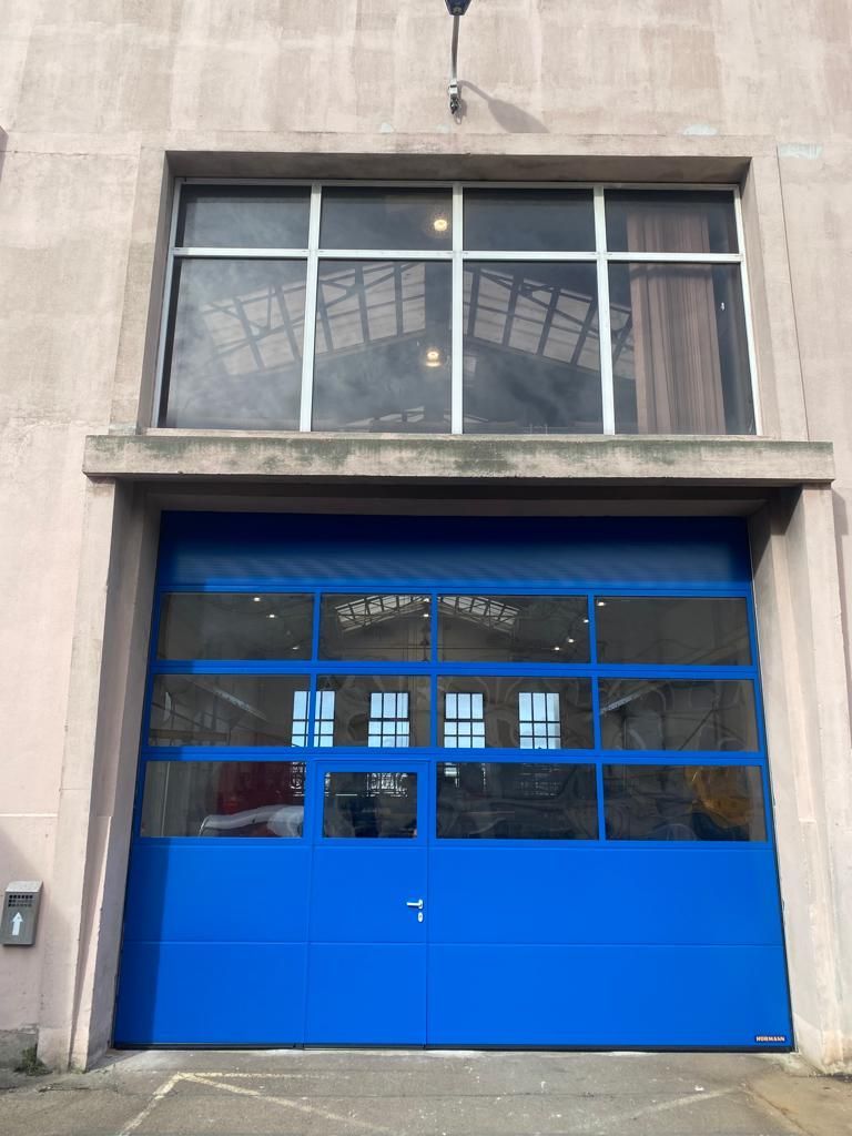 Porte de garage industrielle bleue sous une grande façade vitrée d'un bâtiment en béton