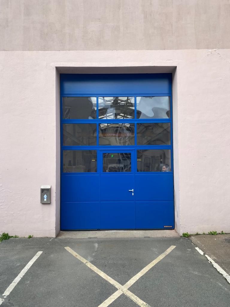 Porte de garage bleue vitrée dans un mur beige, avec une place de parking délimitée par des lignes blanches en forme de X devant.