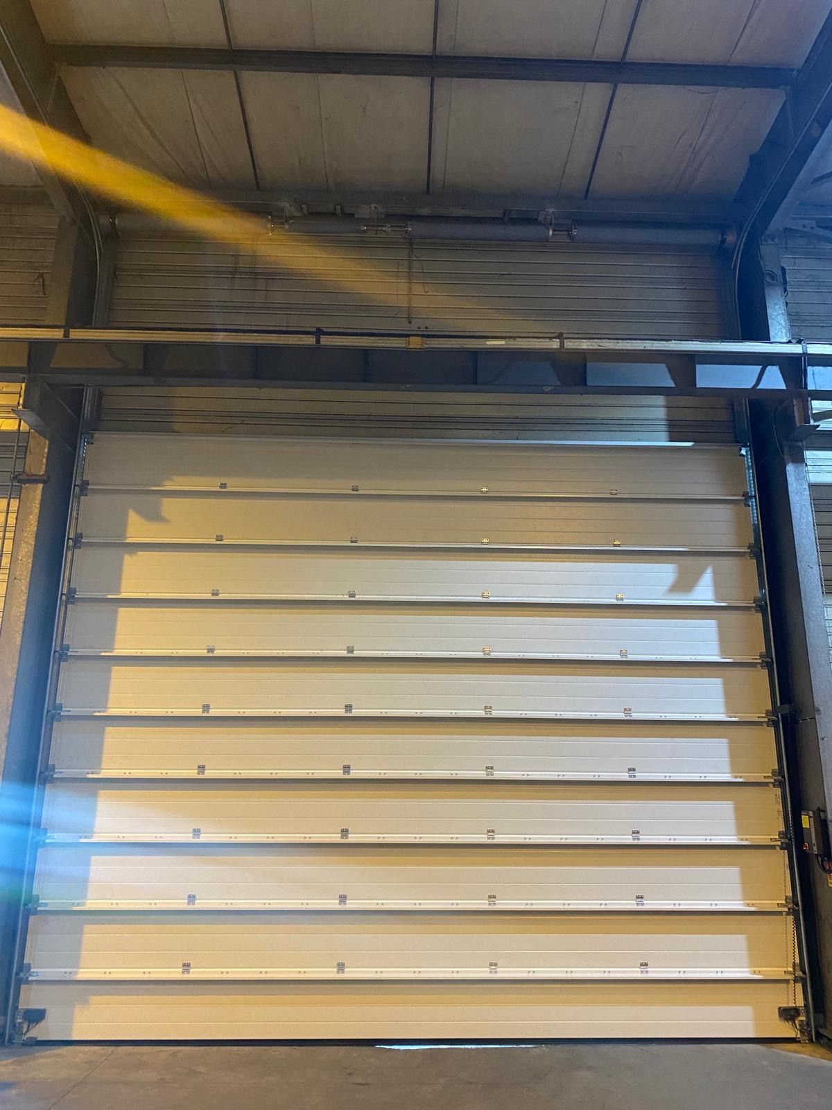 Porte de garage sectionnelle blanche à l'intérieur d'un entrepôt industriel, avec des poutres métalliques et une sangle jaune au plafond.