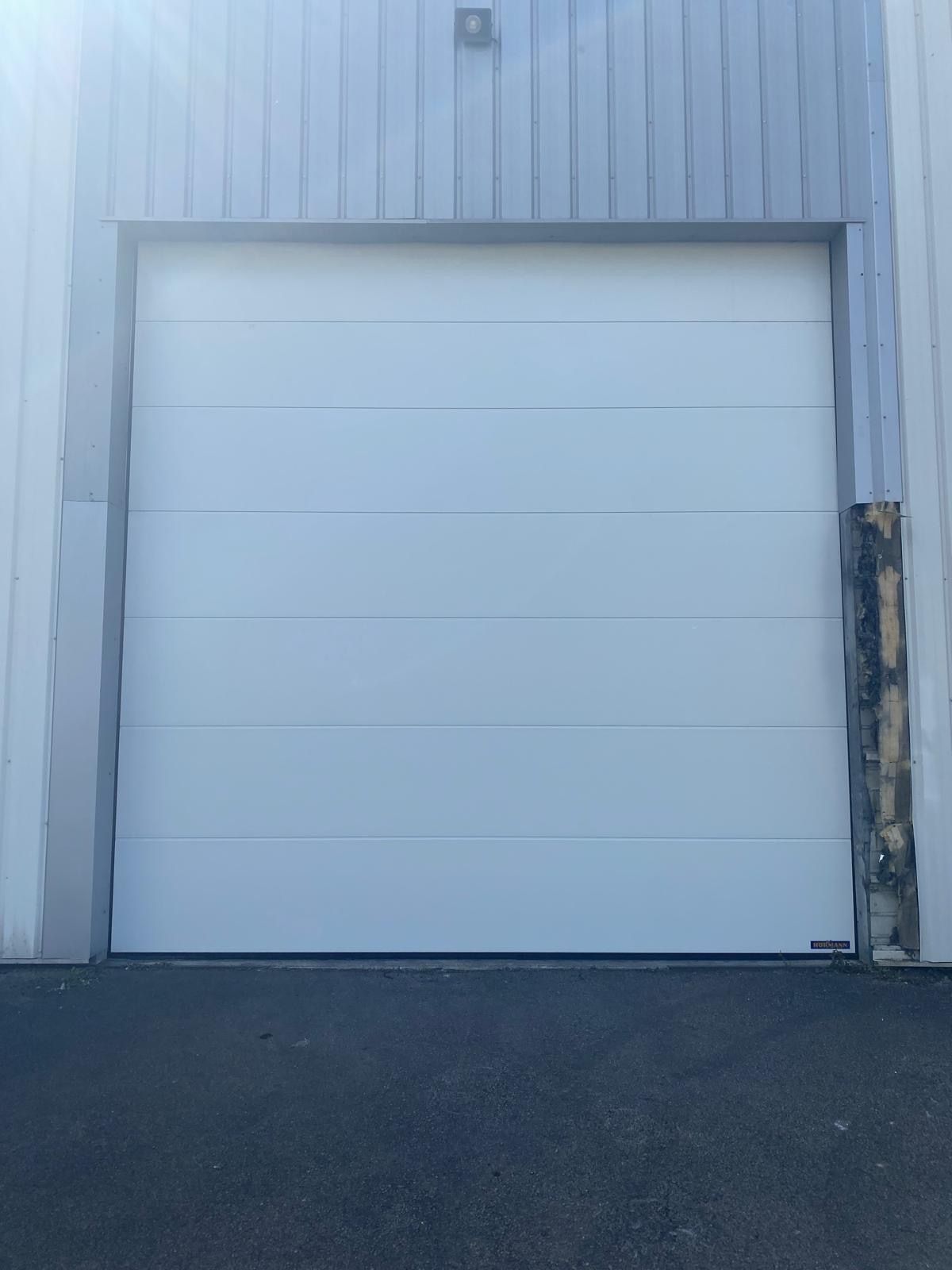 Porte de garage blanche sur un bâtiment à façade métallique, avec un premier plan en asphalte foncé.