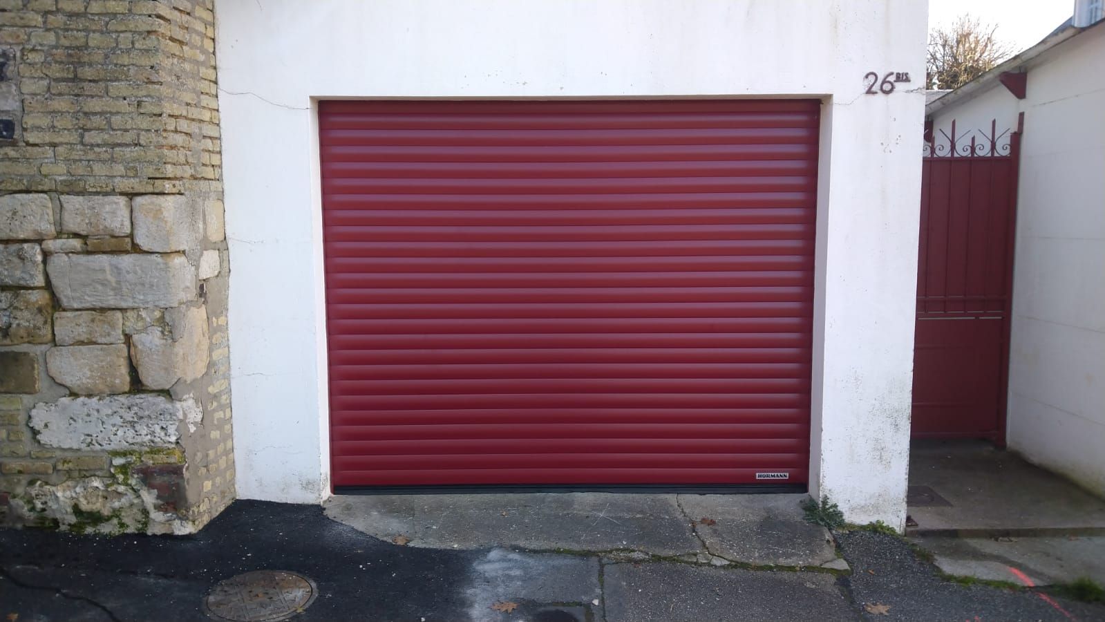 Porte de garage sectionnelle en métal rouge dans une façade d'immeuble en pierre blanche avec une porte latérale étroite.