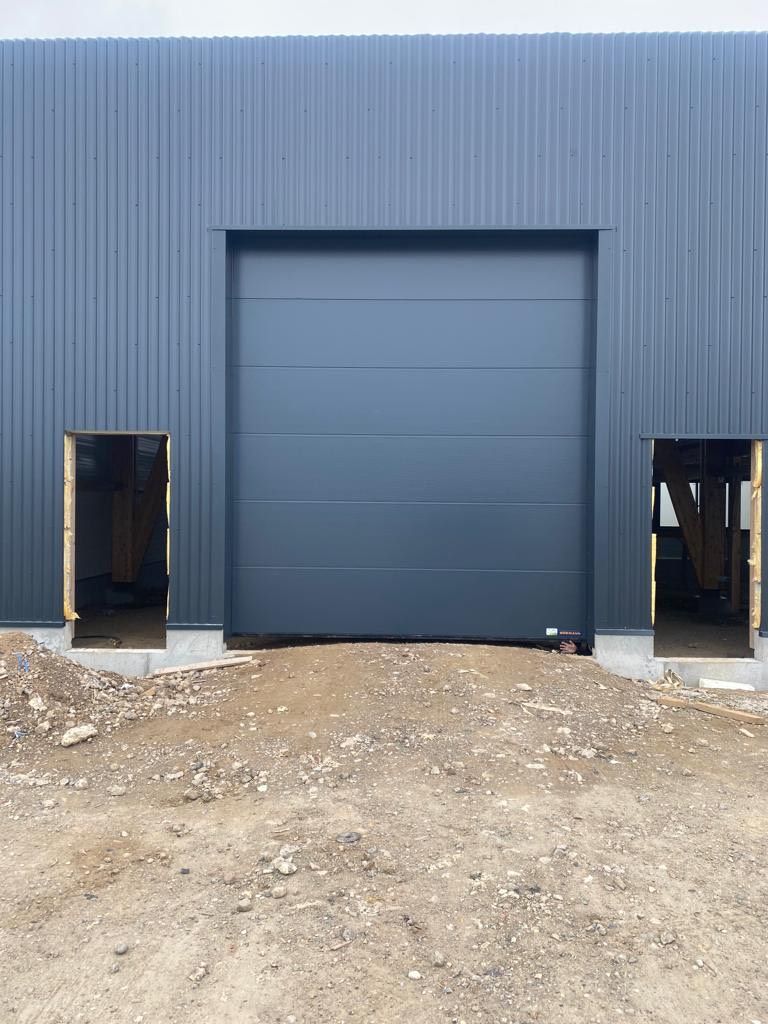 Bâtiment industriel bleu avec une grande porte sectionnelle et deux petites entrées latérales sur un terrain gravillonné
