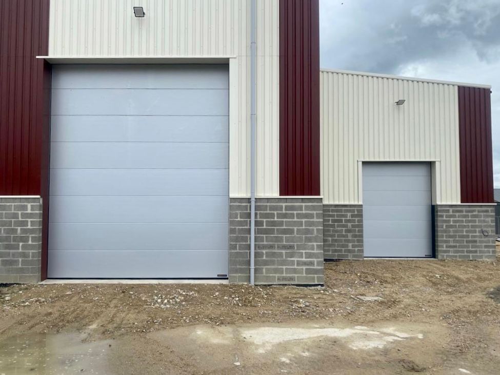 Deux portes de garage enroulables grises sur un bâtiment industriel métallique avec des garnitures verticales rouges et une base en parpaings.