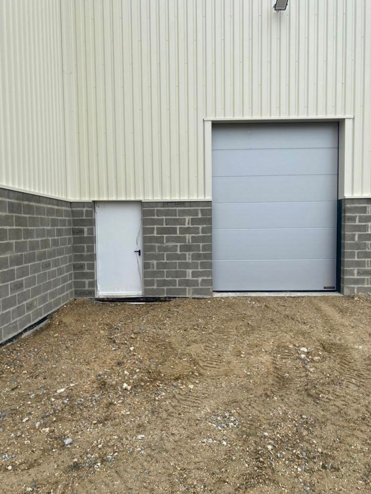 Extérieur d'un bâtiment industriel avec un bardage gris, une petite porte blanche et une grande porte de garage sectionnelle grise sur un sol en gravier.